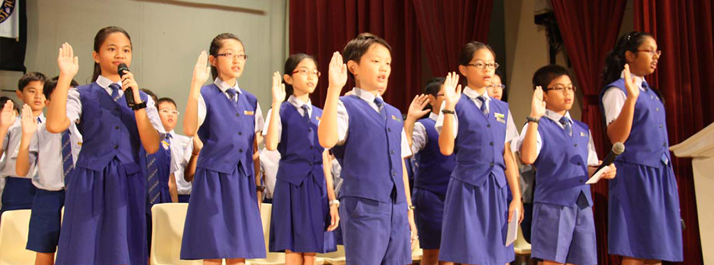 Students in blue uniforms raise their right hands on a stage during what appears to be a pledge ceremony.